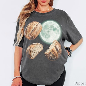 May include: A charcoal gray short-sleeve t-shirt featuring a graphic print of three sourdough bread loaves arranged around a glowing full moon. The design includes a whole loaf, a sliced loaf, and a partially eaten loaf against a dark background.