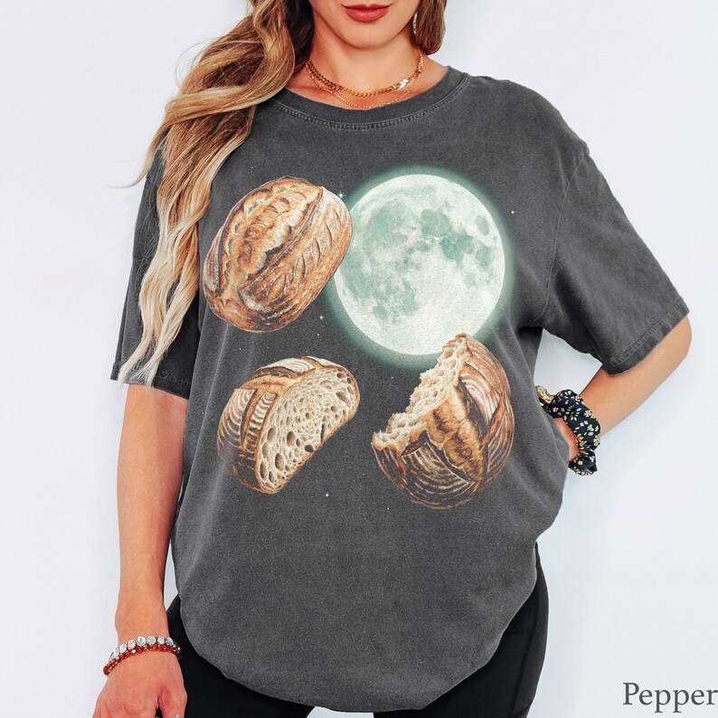 May include: A charcoal gray short-sleeve t-shirt featuring a graphic print of three sourdough bread loaves arranged around a glowing full moon. The design includes a whole loaf, a sliced loaf, and a partially eaten loaf against a dark background.
