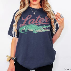 May include: A navy blue short-sleeve t-shirt featuring a graphic of a green alligator positioned below the word "Later" written in a red script font with a white outline. The shirt has a crew neckline and a relaxed, loose-fitting silhouette.