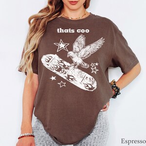 May include: An espresso brown short-sleeve t-shirt featuring a white graphic print of a bird flying above a skateboard with flame designs and stars. The text "thats coo" is printed in white above the bird. The shirt has a crew neckline.