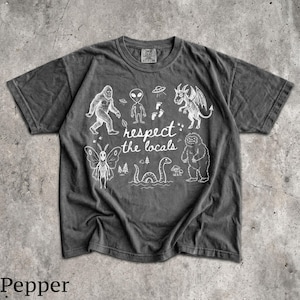 May include: A charcoal gray short-sleeve t-shirt featuring white line art illustrations of cryptids and the text "respect the locals." The graphic includes a moth-like creature, a bigfoot, an alien, a dragon, and a sea serpent among trees.