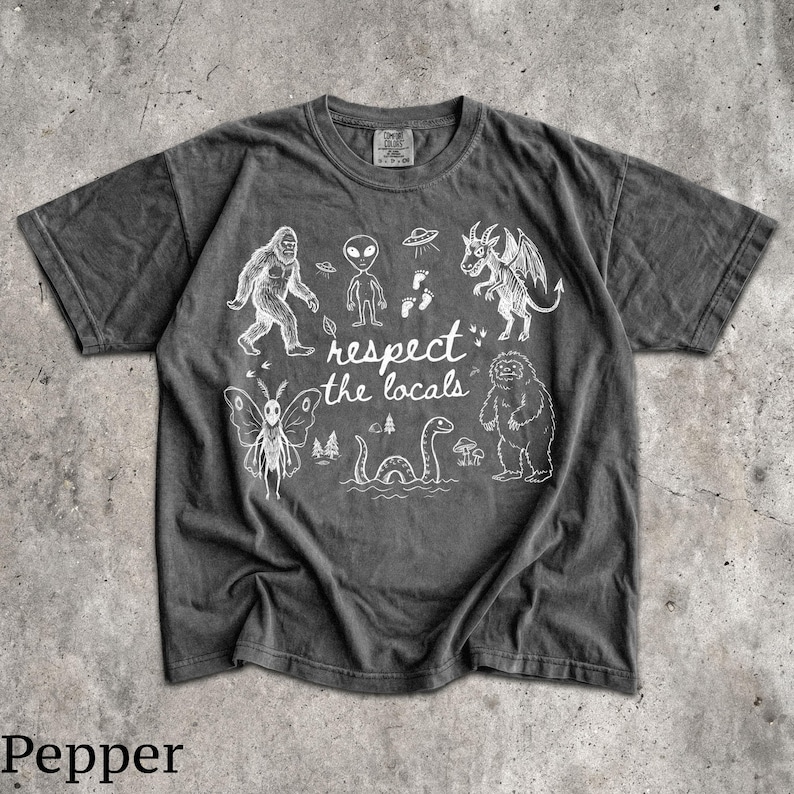 May include: A charcoal gray short-sleeve t-shirt featuring white line art illustrations of cryptids and the text "respect the locals." The graphic includes a moth-like creature, a bigfoot, an alien, a dragon, and a sea serpent among trees.