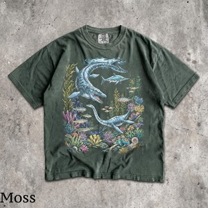Op de afbeelding: Mosgroen t-shirt met een gedetailleerde illustratie van prehistorische zeereptielen die zwemmen tussen kleurrijk koraal en vissen. Het ontwerp bevat een grote mosasaurus en een plesiosaurus. Het woord "Moss" staat onderaan gedrukt.