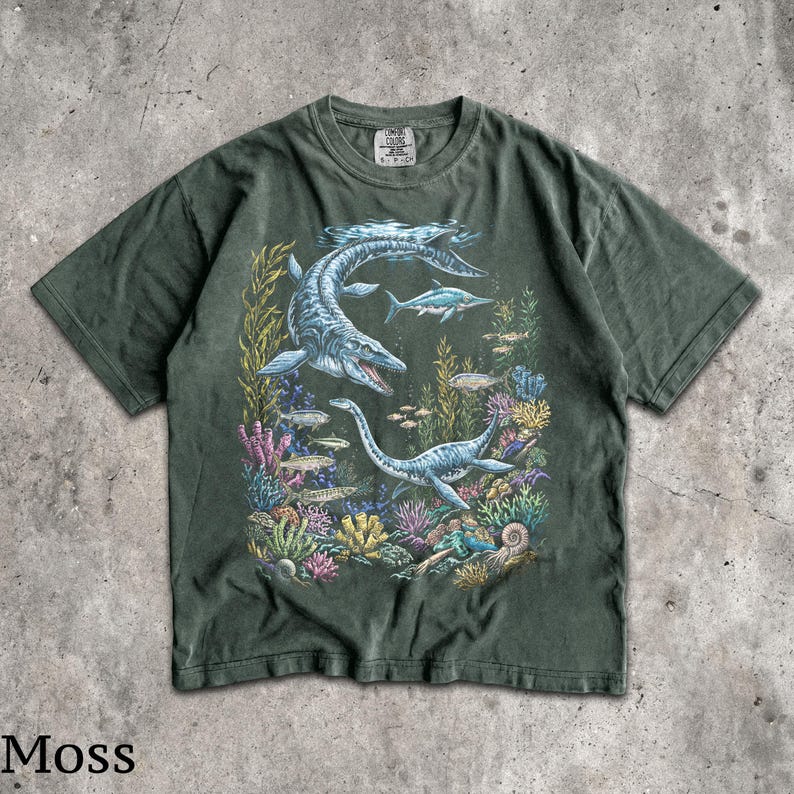 Op de afbeelding: Mosgroen t-shirt met een gedetailleerde illustratie van prehistorische zeereptielen die zwemmen tussen kleurrijk koraal en vissen. Het ontwerp bevat een grote mosasaurus en een plesiosaurus. Het woord "Moss" staat onderaan gedrukt.