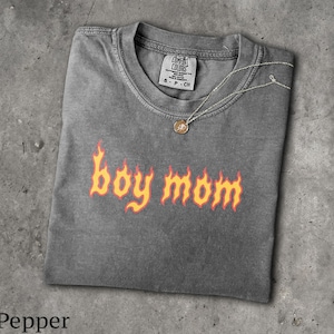 Könnte beinhalten: Ein anthrazitfarbenes Kurzarm-T-Shirt mit Rundhalsausschnitt und einem Comfort Colors Markenetikett. Auf der Vorderseite befindet sich der Schriftzug "boy mom" in einer stilisierten orange-gelben Flammenschrift. Eine dünne silberfarbene Kette liegt auf dem Kragen.