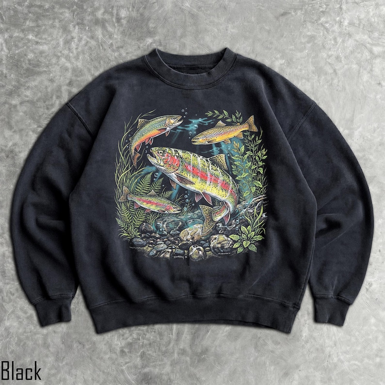 May include: A black crewneck sweatshirt featuring a detailed graphic print of three rainbow trout swimming among green aquatic plants and rocks. The central fish is large and colorful, while two smaller trout are positioned above and below it.