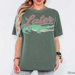 May include: A moss green short-sleeve t-shirt featuring a graphic of a green alligator positioned below the word Later written in a red cursive font. The shirt has a crew neckline and a relaxed fit, showcasing a screen-printed design on the front.