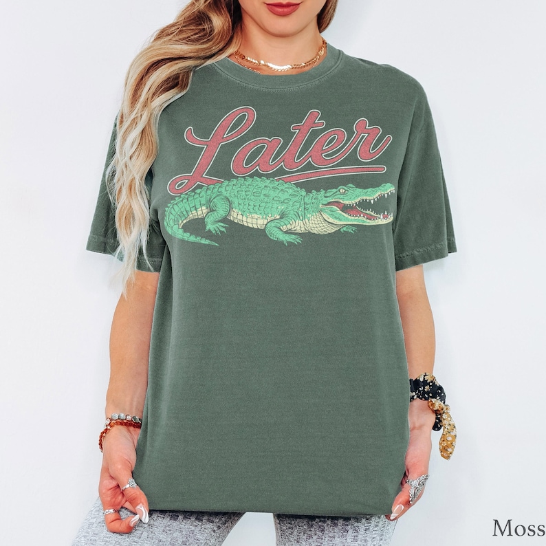 May include: A moss green short-sleeve t-shirt featuring a graphic of a green alligator positioned below the word Later written in a red cursive font. The shirt has a crew neckline and a relaxed fit, showcasing a screen-printed design on the front.