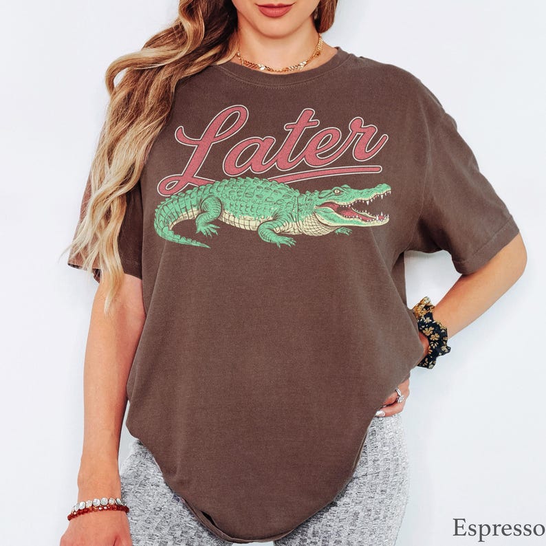 May include: A dark brown short-sleeve t-shirt featuring the word Later in a cursive red font above a graphic illustration of a green alligator with an open mouth. The shirt has a crew neckline and a relaxed fit, showcasing a casual design.