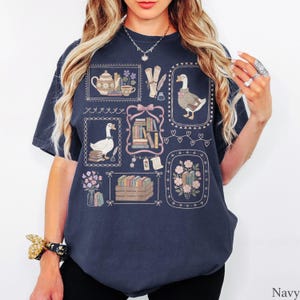 Puede incluir: Una camiseta azul marino con un estampado gr&aacute;fico de varias ilustraciones enmarcadas que incluyen gansos con gafas, pilas de libros, una tetera, una taza de t&eacute;, flores y pergaminos. El dise&ntilde;o est&aacute; dispuesto en un patr&oacute;n de cuadr&iacute;cula.