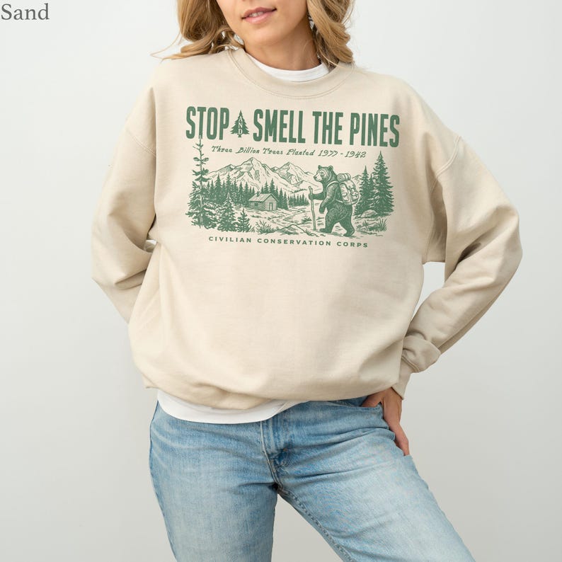 Pode incluir: Uma camisola de gola redonda cor de areia com uma impress&atilde;o gr&aacute;fica verde de um urso a caminhar numa floresta com montanhas e uma cabana. O texto diz STOP SMELL THE PINES, Three Billion Trees Planted 1977-1942, CIVILIAN CONSERVATION CORPS.