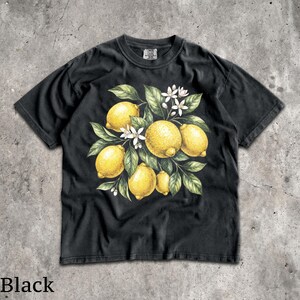 Może przedstawiać: Czarny t-shirt z akwarelową ilustracją cytryn, liści i białych kwiat&oacute;w. Projekt przedstawia ż&oacute;łte cytryny z zielonymi liśćmi i białymi kwiatami, tworząc letni, botaniczny motyw.