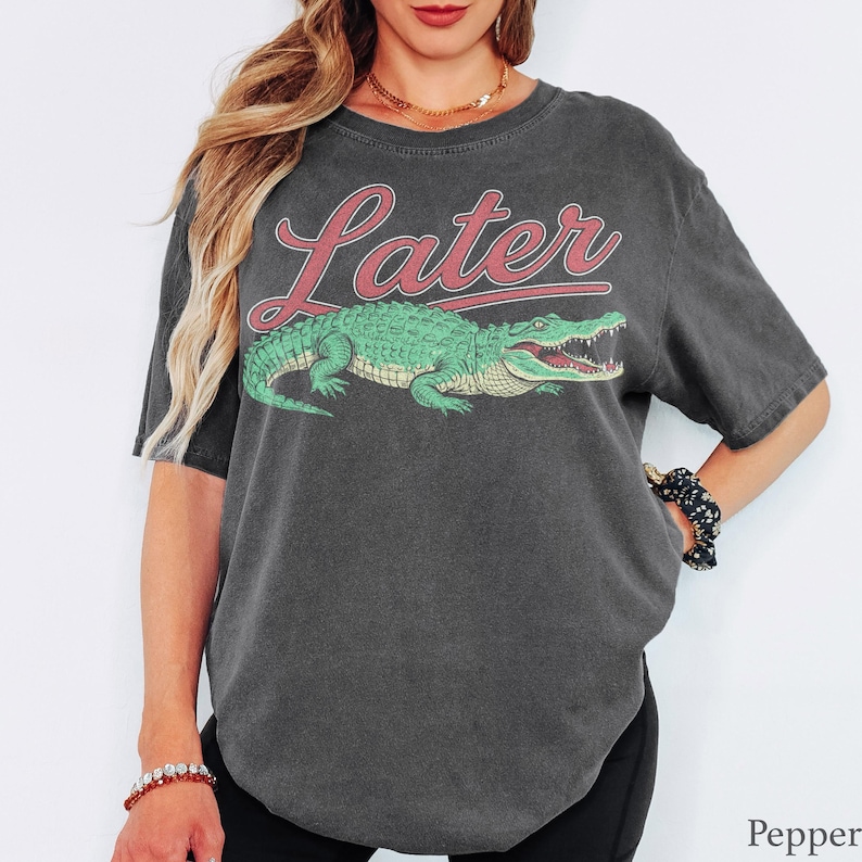 May include: A charcoal grey short-sleeve t-shirt featuring a graphic of a green alligator positioned below the word Later written in a red script font. The shirt has a relaxed fit and a crew neckline, showcasing a vintage-style washed fabric texture.