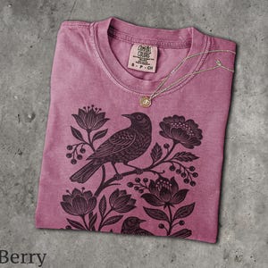 May include: A berry-colored short-sleeve t-shirt featuring a black graphic print of a bird perched on a flowering branch. The shirt has a crew neckline and a Comfort Colors brand label at the inner collar. A thin gold chain necklace rests on the fabric.