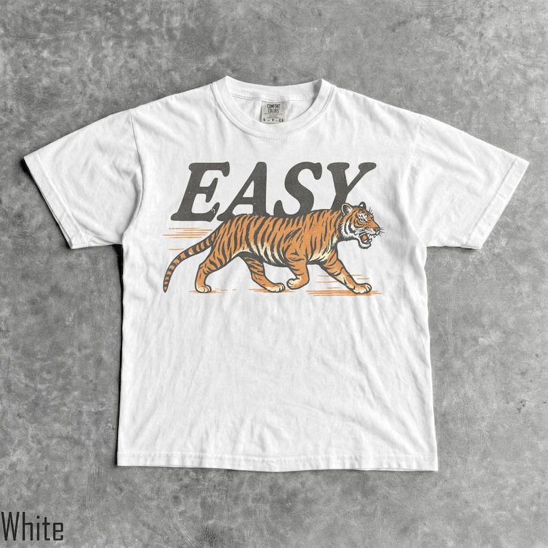 May include: A white short-sleeve t-shirt featuring a graphic of an orange and black tiger walking. Above the tiger, the word EASY is printed in a large, dark gray, distressed font. The shirt has a crew neckline and a standard fit design.