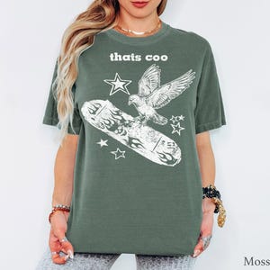 May include: A moss green short-sleeve t-shirt featuring a white graphic print of a bird flying above a skateboard with flame designs. The text "thats coo" is printed in white above the bird, accompanied by several small star shapes.