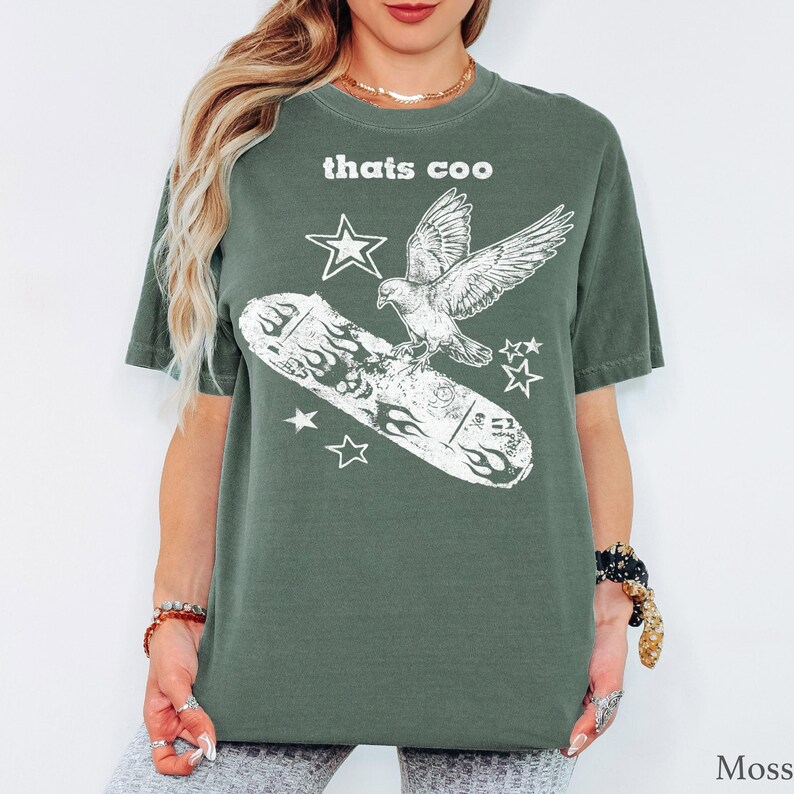 May include: A moss green short-sleeve t-shirt featuring a white graphic print of a bird flying above a skateboard with flame designs. The text "thats coo" is printed in white above the bird, accompanied by several small star shapes.