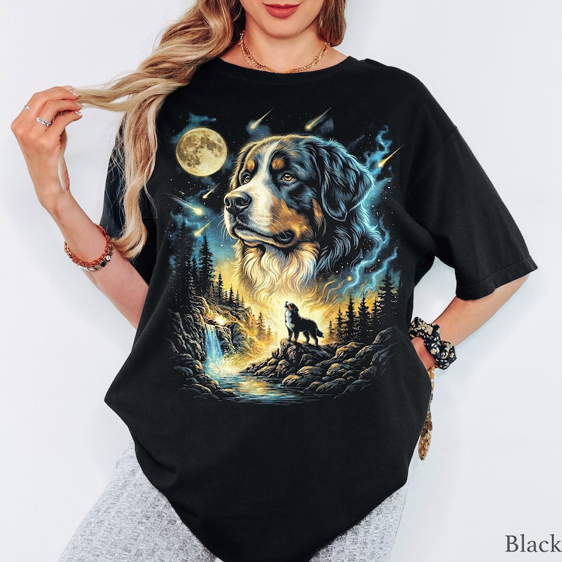 May include: A black short-sleeved t-shirt featuring a graphic print of a Bernese Mountain Dog. The design includes a large dog portrait, a smaller dog standing on a rocky cliff near a waterfall, a full moon, and falling stars in a night sky.