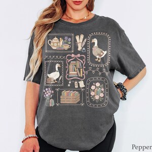 Puede incluir: Una camiseta gris carb&oacute;n de manga corta con un estampado gr&aacute;fico de varias ilustraciones enmarcadas que incluyen gansos, pilas de libros, una tetera, pergaminos y arreglos florales en tonos pastel sobre la tela oscura.