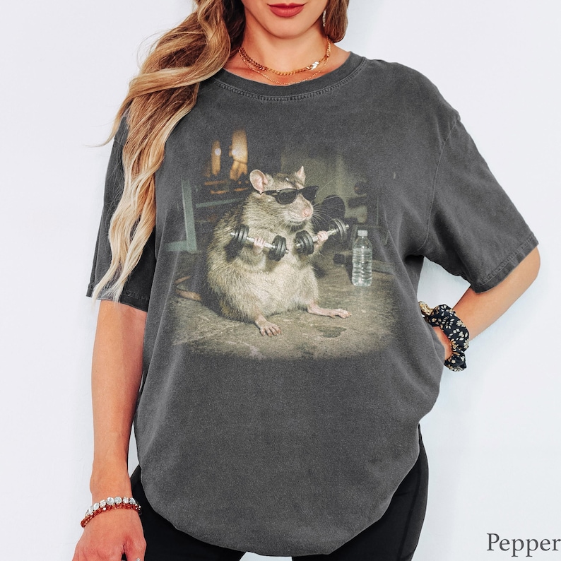 May include: A charcoal gray short-sleeve t-shirt featuring a graphic print of a rat wearing black sunglasses and lifting small dumbbells in a gym setting, with a clear water bottle visible on the floor next to the rat and the word Pepper.