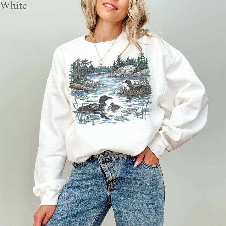 May include: A white long-sleeve sweatshirt featuring a graphic illustration of a lake scene with two loons, one small chick, a nest, pine trees, and rocky terrain. The design is centered on the chest, showcasing natural colors like green, blue, and brown.