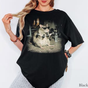 May include: A black t-shirt featuring a graphic print of a rat wearing black sunglasses while lifting small dumbbells in a gym setting. A clear plastic water bottle is positioned on the floor next to the rat in the center of the shirt design.
