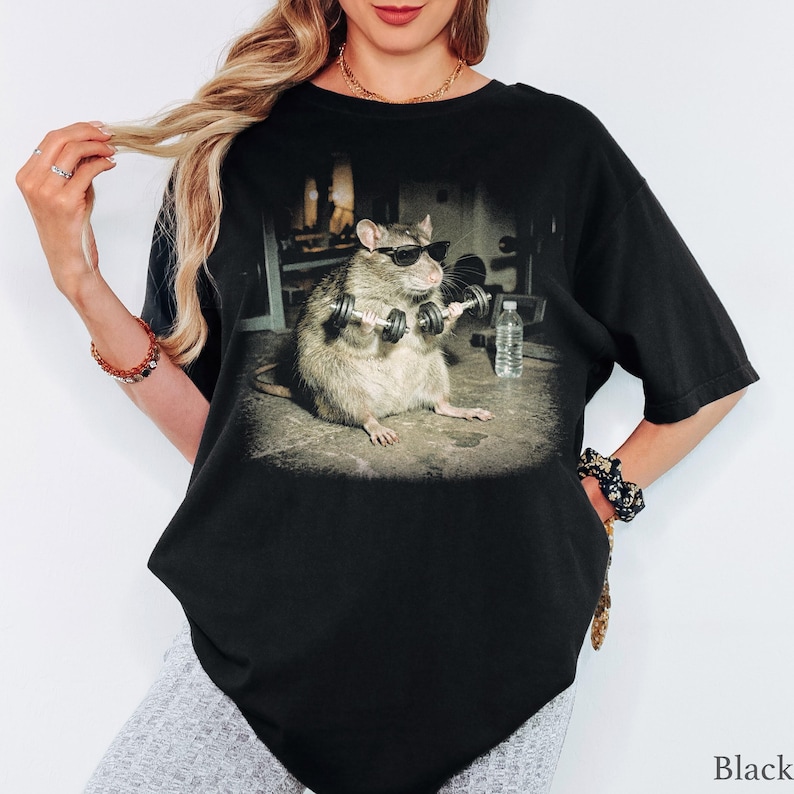 May include: A black t-shirt featuring a graphic print of a rat wearing black sunglasses while lifting small dumbbells in a gym setting. A clear plastic water bottle is positioned on the floor next to the rat in the center of the shirt design.