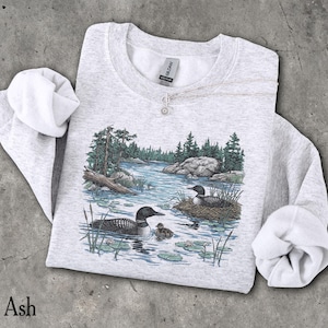 May include: An ash gray crewneck sweatshirt featuring a detailed graphic print of two loons swimming in a lake with a small chick, surrounded by pine trees, rocks, and lily pads. The garment has long sleeves and a ribbed collar with a Gildan brand tag.