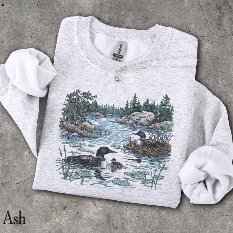 May include: An ash gray crewneck sweatshirt featuring a detailed graphic print of two loons swimming in a lake with a small chick, surrounded by pine trees, rocks, and lily pads. The garment has long sleeves and a ribbed collar with a Gildan brand tag.