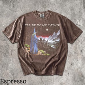 Op de afbeelding: Een bruin t-shirt met korte mouwen en een grafische print van een tovenaar in een blauwe mantel die op een klif staat en uitkijkt over een berglandschap en een kasteel. De tekst "I'LL BE IN MY OFFICE" staat in het wit boven de afbeelding.