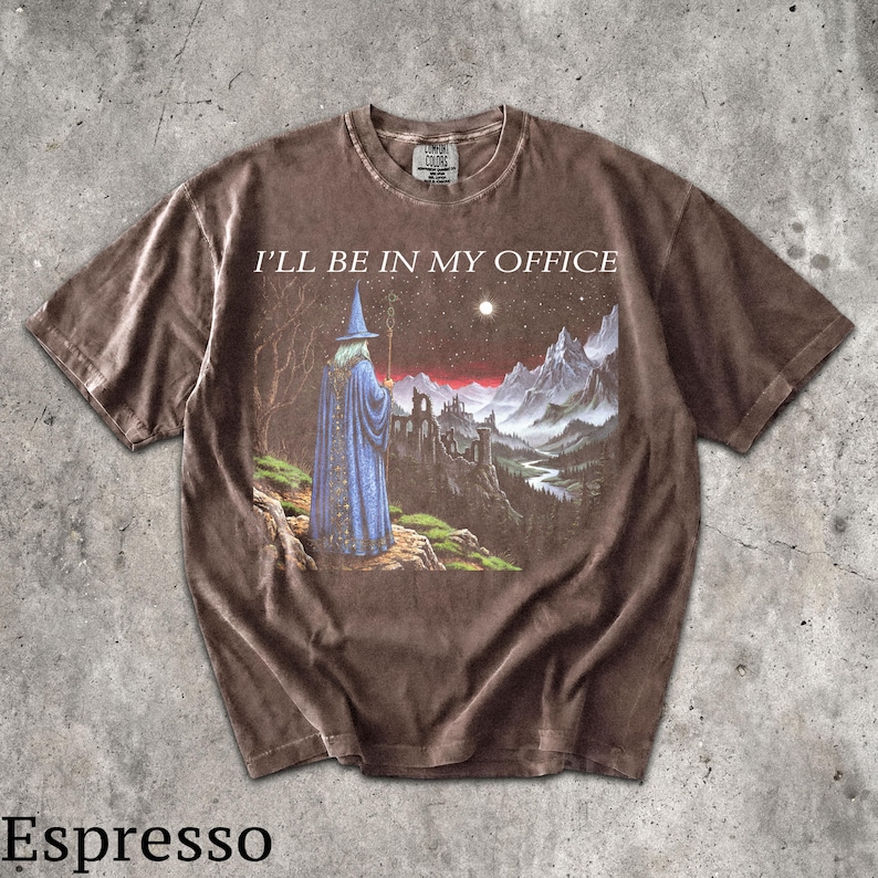 Op de afbeelding: Een bruin t-shirt met korte mouwen en een grafische print van een tovenaar in een blauwe mantel die op een klif staat en uitkijkt over een berglandschap en een kasteel. De tekst "I'LL BE IN MY OFFICE" staat in het wit boven de afbeelding.