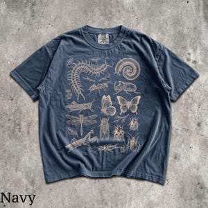 May include: A navy blue short-sleeve t-shirt featuring a detailed, light-colored graphic print of various insects, including a centipede, butterfly, dragonfly, beetle, and mantis, arranged across the front with a visible neck label.
