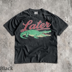 Puede incluir: Camiseta negra con un dise&ntilde;o de cocodrilo verde y la palabra "Later" en letras rojas. El cocodrilo tiene la boca abierta. La camiseta es de cuello redondo y est&aacute; hecha de un material suave.