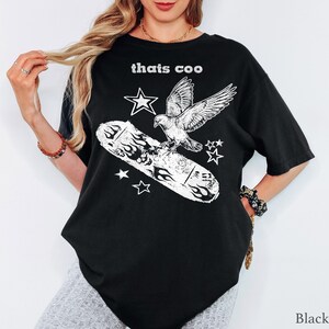 May include: A black short-sleeve t-shirt featuring a white graphic print of a pigeon perched on a skateboard with flames. The text "thats coo" is printed in white above the graphic, which is surrounded by several white star shapes.