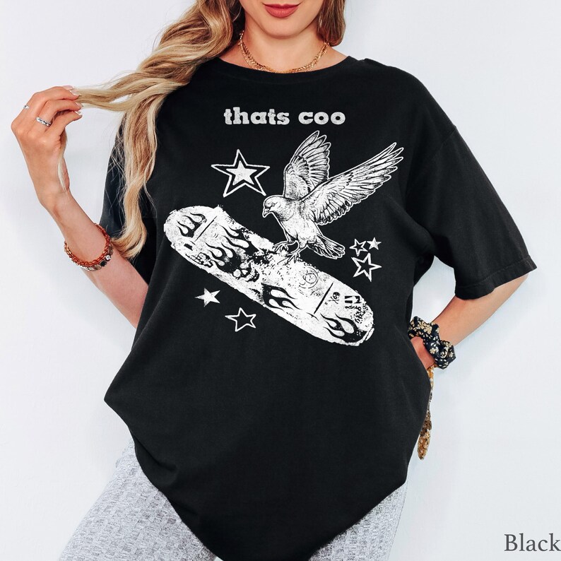 May include: A black short-sleeve t-shirt featuring a white graphic print of a pigeon perched on a skateboard with flames. The text "thats coo" is printed in white above the graphic, which is surrounded by several white star shapes.