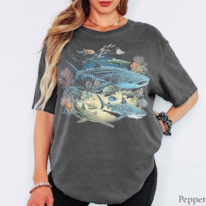 May include: A charcoal gray short-sleeve t-shirt featuring a detailed graphic print of various marine life, including a large whale shark, a sawfish, and a smaller shark swimming among coral reefs and schools of fish. The fabric has a washed-out look.