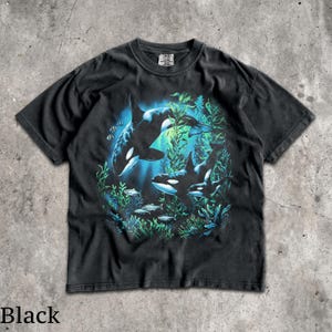 May include: A black short-sleeve t-shirt featuring a graphic print of two orcas swimming among green seaweed and small fish. The design uses shades of blue and green to depict an underwater scene, with the Comfort Colors brand tag at the neck.