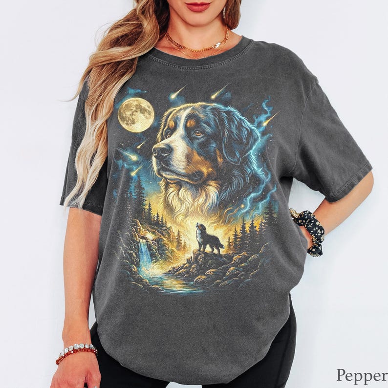 May include: A charcoal gray short-sleeve t-shirt featuring a graphic print of a Bernese Mountain Dog head above a smaller dog standing on a rocky cliff near a waterfall and forest, all set against a night sky with a full moon and shooting stars.