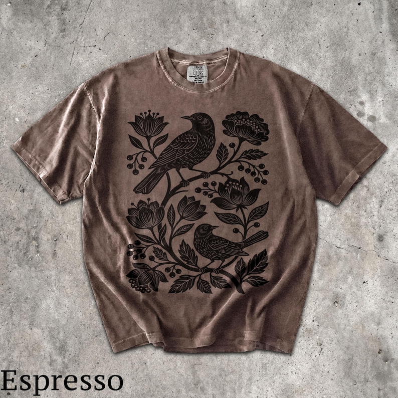 May include: A brown short-sleeve t-shirt featuring a black graphic print of two birds perched among stylized flowers and berries. The shirt has a crew neckline and a label at the inner collar that reads Comfort Colors. The fabric has a washed appearance.