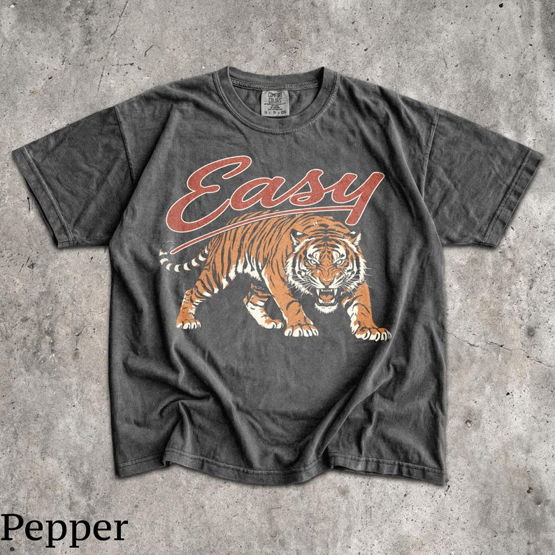 May include: A charcoal grey short-sleeve t-shirt featuring a graphic of a prowling tiger in orange and black tones. Above the tiger, the word "Easy" is printed in a stylized red script font. The shirt has a crew neckline and a faded, worn-in texture.