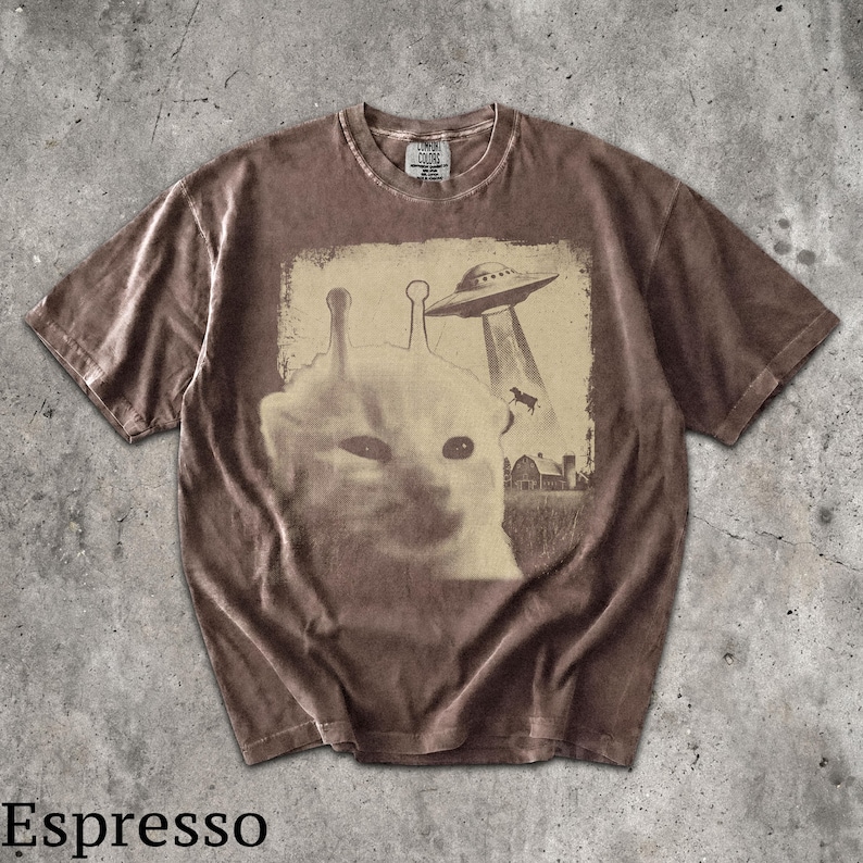 May include: A brown short-sleeve t-shirt featuring a graphic print of a cat with antennae, a flying saucer beaming down a cow, and a barn. The shirt has a crew neckline and a label at the inner collar that reads Comfort Colors. The color is espresso.