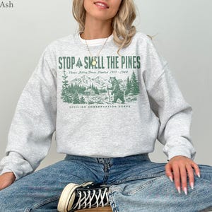 Pode incluir: Uma camisola cinzenta clara de gola redonda com uma impress&atilde;o gr&aacute;fica verde de um urso numa floresta com montanhas e o texto STOP SMELL THE PINES, Three Billion Trees Planted 1933-1942, e CIVILIAN CONSERVATION CORPS na parte frontal.