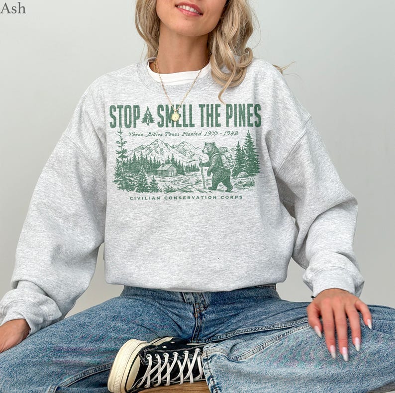 Pode incluir: Uma camisola cinzenta clara de gola redonda com uma impress&atilde;o gr&aacute;fica verde de um urso numa floresta com montanhas e o texto STOP SMELL THE PINES, Three Billion Trees Planted 1933-1942, e CIVILIAN CONSERVATION CORPS na parte frontal.