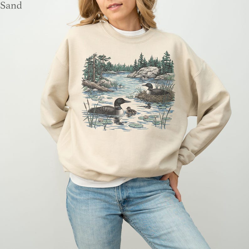 May include: A sand-colored crewneck sweatshirt featuring a graphic illustration of two loons swimming in a lake with a small chick, surrounded by pine trees, rocks, and lily pads. The text "Sand" is printed in the upper left corner of the image.