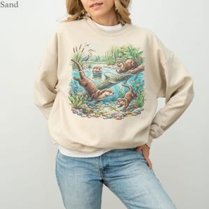 Op de afbeelding: Een zandkleurige sweater met ronde hals en een kleurrijke grafische print van otters die in een rivier zwemmen met waterplanten, rotsen en een reiger. Het woord Sand staat linksboven in de afbeelding boven de sweater.