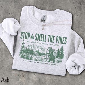 Pode incluir: Uma camisola de gola redonda em cinzento mesclado claro com uma impress&atilde;o gr&aacute;fica verde-escura. O design mostra um urso com uma mochila numa paisagem florestal com montanhas, o texto STOP AND SMELL THE PINES, Three Billion Trees Planted 1977-1942 e CIVILIAN CONSERVATION CORPS.