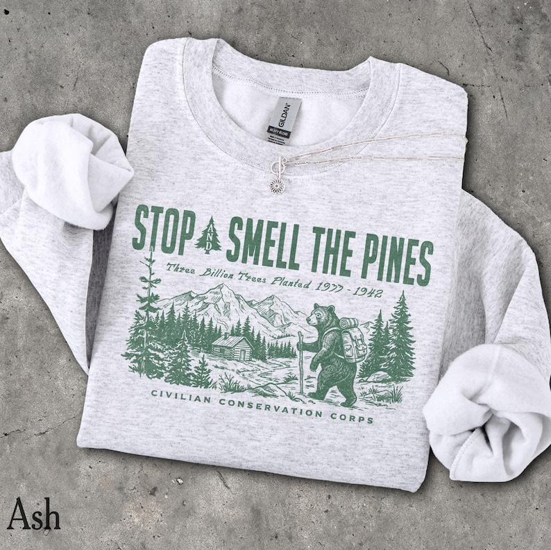 Pode incluir: Uma camisola de gola redonda em cinzento mesclado claro com uma impress&atilde;o gr&aacute;fica verde-escura. O design mostra um urso com uma mochila numa paisagem florestal com montanhas, o texto STOP AND SMELL THE PINES, Three Billion Trees Planted 1977-1942 e CIVILIAN CONSERVATION CORPS.