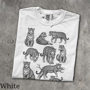 May include: A white short-sleeve t-shirt featuring a black ink-style graphic print of multiple jaguars in various poses. The shirt has a crew neckline and a Comfort Colors brand label printed on the inside of the collar area.