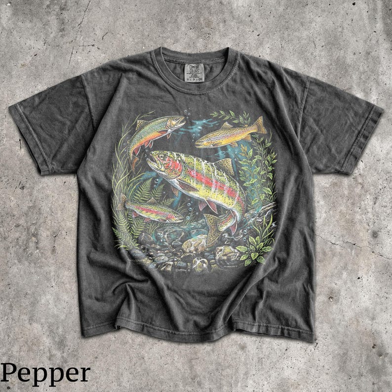 May include: Dark gray t-shirt featuring a colorful illustration of several fish swimming in a freshwater environment. The fish are surrounded by aquatic plants and rocks. The word "Pepper" is printed at the bottom.