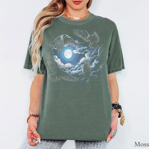 May include: A moss green short-sleeve t-shirt featuring a graphic print of four black ravens flying around a bright full moon surrounded by white clouds. The design is centered on the chest of the shirt, which has a crew neckline and a relaxed fit.